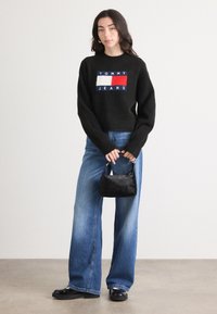 Svart genser med ribbestruktur med "TOMMY JEANS"-logo i blått, rødt og hvitt. Vide blå jeans og en liten svart håndveske.