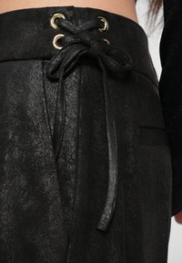 Black trousers featuring a textured finish, secured with a black fabric tie through gold eyelets at the waist, with a side pocket visible.