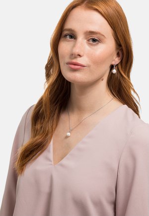 Silberne Halskette mit einem runden Perlenanhänger und passenden Perlenohrhängern. Bluse mit V-Ausschnitt in hellem Rosa. Glatte Stofftextur.