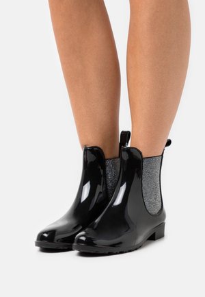 Bottes de pluie noires brillantes à talon, avec des panneaux latéraux élastiques gris, portées sur des jambes nues sur un fond blanc.