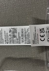 Etykieta z instrukcjami pielęgnacji nadrukowana na białej tkaninie, z czarnym tekstem, symbolami i logo. Tło to teksturowana szara tkanina.