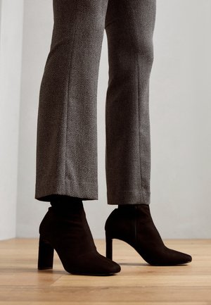 Vue de la longueur des jambes d'une personne portant un pantalon gris à motif chevrons et des bottines noires en daim à talons hauts, debout sur un sol en bois clair.