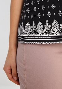 Top nero con intricati motivi bianchi e un orlo decorativo. Il tessuto è leggero, abbinato a pantaloni rosa chiaro.