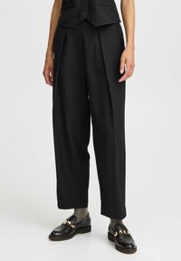 Svarta skräddarsydda högmidjade byxor med veck, slät textur, vida ben, i kombination med svarta loafers och mönstrade strumpor.