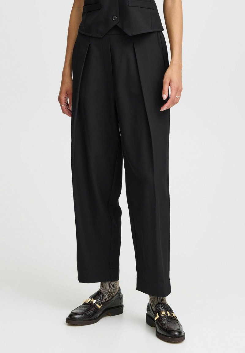 Svarta skräddarsydda högmidjade byxor med veck, slät textur, vida ben, i kombination med svarta loafers och mönstrade strumpor.