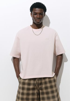 Jeune homme portant un t-shirt oversize rose clair, un collier chaîne en argent et un pantalon marron à carreaux, les mains dans les poches, debout devant un fond blanc.