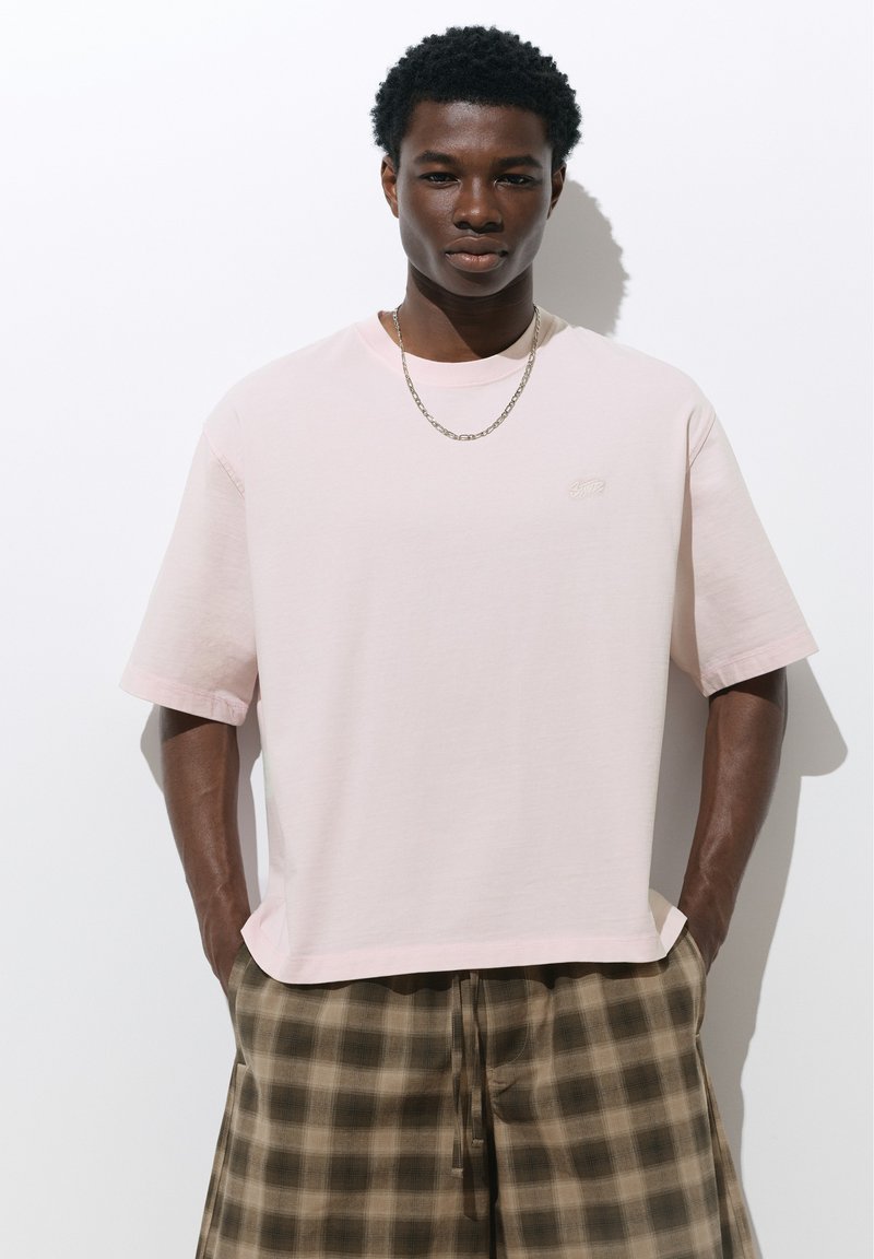 Jeune homme portant un t-shirt oversize rose clair, un collier chaîne en argent et un pantalon marron à carreaux, les mains dans les poches, debout devant un fond blanc.