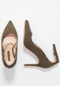 Olivově zelené semišové boty na vysokém podpatku s špičatou špičkou a otevřenými stranami. Obsahují pásek na kotníku s kovovými prvky a béžovou stélku.