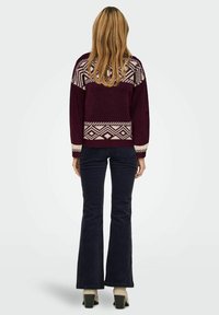 Mørkerød strikket sweater med hvid geometrisk mønster på skuldrene og kantene, parret med mørke flared jeans og beige blokhæl støvler.
