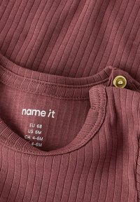 Tissu côtelé bordeaux avec un col rond, présentant une fermeture à boutons pression et des étiquettes de soin affichant la taille et les informations sur la marque.