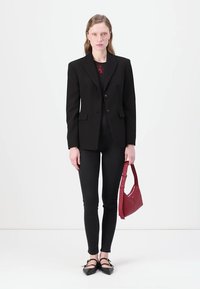 Črni elegantni blazer z dvema gumboma, v kombinaciji s pričvrščenimi črnimi kavbojkami. Model drži rdečo usnjeno torbico in nosi črne ravne čevlje.