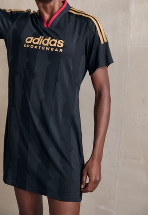 Robe de sport noire Adidas avec des bandes dorées aux épaules et un logo Adidas doré portée par une personne devant un mur beige uni.