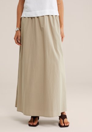 Žena v béžovej maxi sukni, bielom tílku bez rukávov, striebornom náramku a čiernych sandáloch s otvorenou špičkou na neutrálnej background.