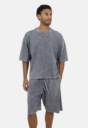 Mężczyzna ubrany w luźny zestaw w kolorze wyblakłej szarości, składający się z oversize'owej koszulki z krótkim rękawem oraz krótkich spodenek do kolan z regulowanym sznurkiem w pasie.