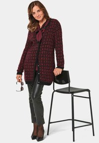 Gestrickter langer Cardigan in Dunkelrot und Schwarz mit einem strukturierten Muster, mit Knopfleiste und gerippten Bündchen. Kombiniert mit engen schwarzen Hosen.