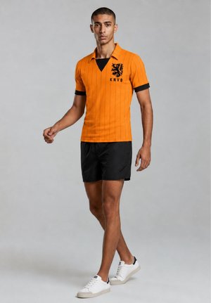 Jonge man loopt, draagt een oranje sportshirt met korte mouwen met zwart leeuwenembleem, zwarte korte broek en witte sneakers op een grijze achtergrond.