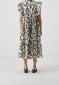 Krémová a černá midi šaty s květinovým potiskem, s nařasenými rukávy, zdůrazněným pasem a detailním vrstveným sukní. Černé boty na blokovém podpatku.