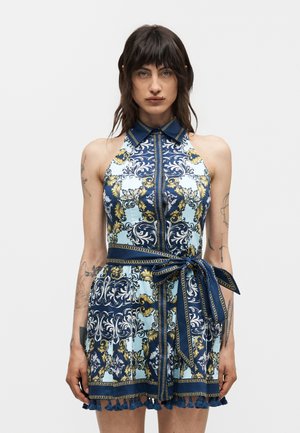 Γυναίκα με σκουρόχρωμα μαλλιά που φοράει μπλε, χρυσό και λευκό φόρεμα χωρίς μανίκια με κολάρο και ζώνη δεμένη στη μέση, με απλό φόντο.