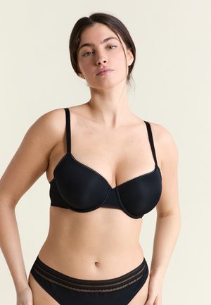 Mujer con sujetador negro con aros y ropa interior a juego, de pie contra un fondo claro y liso, con expresión neutral.