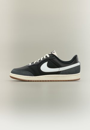Lavtop sort sneaker med hvid Nike-swoosh, hvide snørebånd og beige sål med brun ydersål, vist på neutral baggrund.