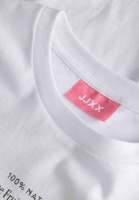 Col blanc de T-shirt en coton avec une étiquette rose portant la mention "JJXX" et un texte partiel indiquant 100 % tissu naturel.