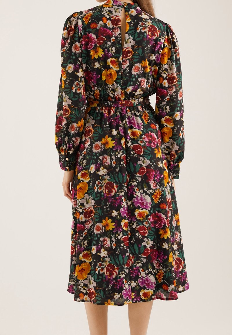 Femme portant une robe midi à manches longues ceinturée avec un motif floral coloré sur fond noir, vue de dos.