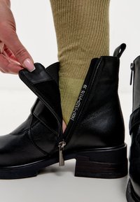 Botín de cuero negro hasta el tobillo con cierre de cremallera, que presenta un acabado texturizado, un tacón estilo madera en contraste y un calcetín ribeteado verde parcialmente visible.