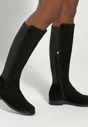 Person wearing knee-high black suede boots with side zipper and elastic panel, standing against a plain light background.