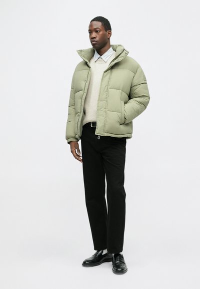 Jasno zielona kurtka puchowa z kapturem, zapinana na zamek błyskawiczny, z dużymi pikowanymi sekcjami, noszona na kremowym swetrze i czarnych spodniach, uzupełniona czarnymi butami.