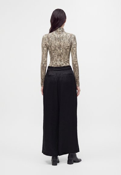 Femme aux longs cheveux foncés portant un haut à manches longues imprimé léopard, un pantalon large noir et des bottines noires, vue de dos.