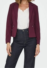 Blazer bordeaux au design ajusté, avec deux poches avant et une texture lisse, associé à un haut blanc à bords festonnés et un jean bleu marine.