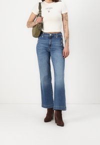 Jeans acampanados de color azul claro con cintura alta, que presentan dos acentos de botón en el lado, combinados con una camiseta blanca ajustada y botas de tacón marrones.