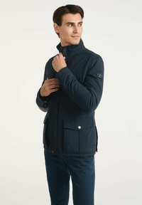 Granatowa kurtka z wysokim kołnierzem, dwoma przednimi kieszeniami i zatrzaskami. Wykonana z gładkiego materiału, z naszywką z logo na rękawie.