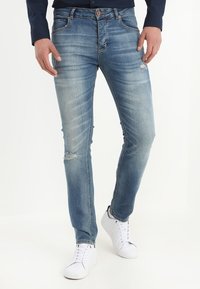Jeans in denim blu chiaro con dettagli strappati, design aderente e gamba dritta. Abbinati a scarpe da ginnastica bianche per un look casual.