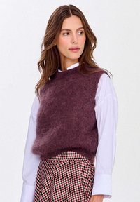 Švestkově barevná, načechraná pletená vesta s kulatým výstřihem, nošená přes bílou košili s límečkem, v kombinaci s kostkovanou sukní.