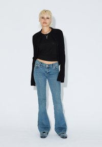 Svart ribbad kofta över en svart kort topp, i kombination med ljusblå utsvängda jeans. Anmärkningsvärda detaljer inkluderar knäppning framtill och en subtil glans.