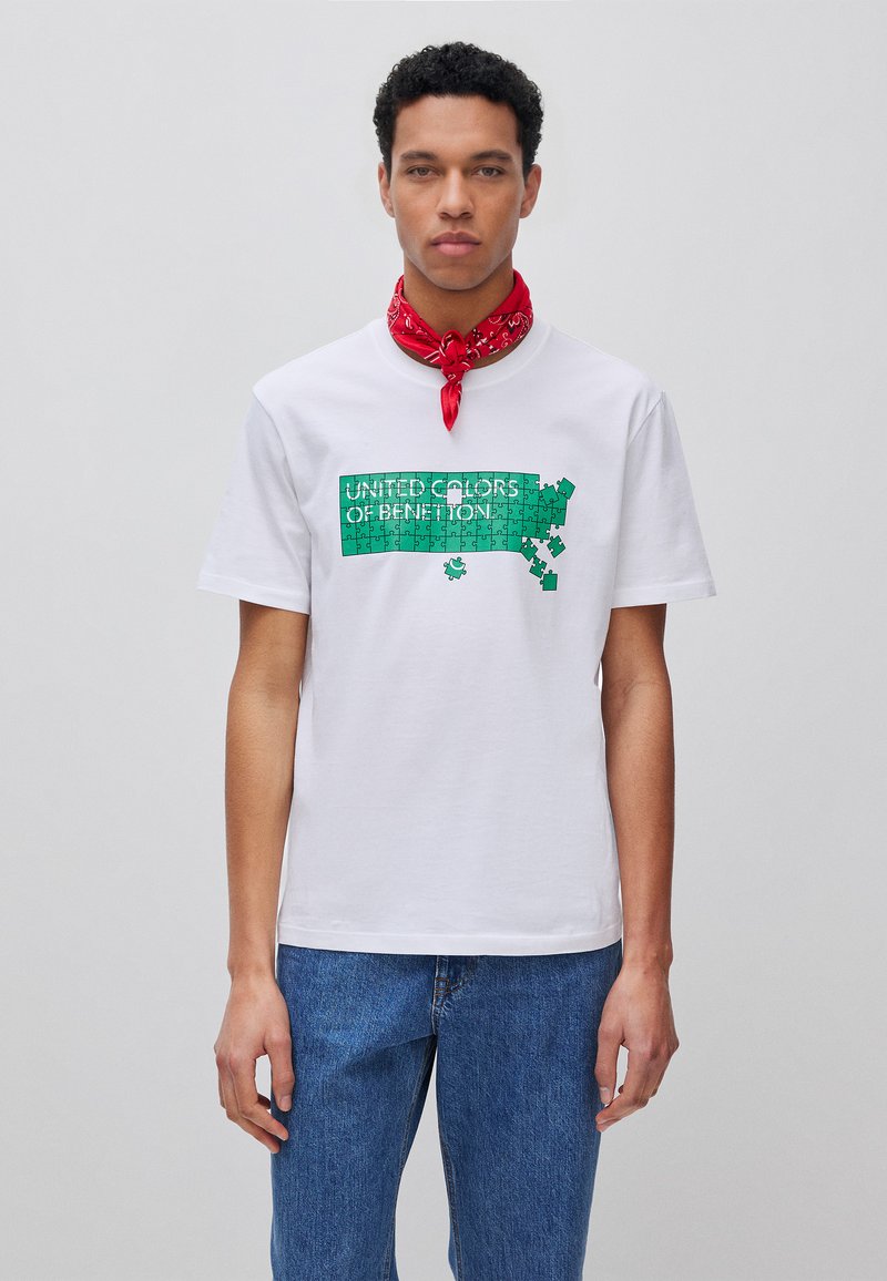 Man draagt een wit T-shirt met een groene puzzelafbeelding en de tekst "United Colors of Benetton" en een rode sjaal, staand tegen een effen achtergrond.