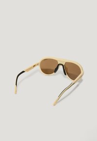 Gafas de sol con una montura de color crema, lentes marrones y detalles en negro; diseño elegante con brazos curvados y puntas de patilla texturizadas.