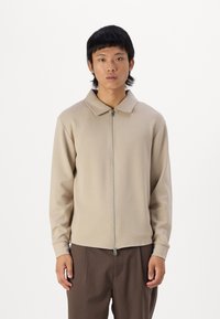 Beige zipzáras dzseki galléros nyakkal, hosszú ujjal és sima textúrával. Barna nadrággal párosítva, a modell egy fehér háttér előtt áll.
