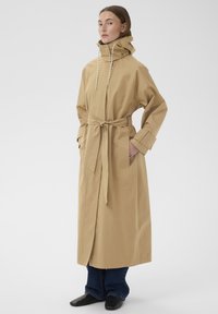 Žena stojící, oblečená v dlouhém béžovém trenčkotě se zavázaným páskem, vysokým límcem se stahovacími šňůrkami, modrých džínách a černých botách na bílém pozadí.