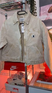 Chaqueta Carhartt beige colgada en una percha negra sobre un estante metálico con cajas de zapatos Nike y Stussy debajo, una zapatilla blanca Nike y una iluminación roja detrás.