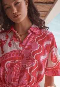 Chemise à motifs rouges et roses avec des dessins nautiques, col à boutons, manches courtes et taille nouée pour une coupe décontractée.