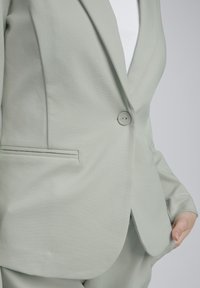 Jasnozielony, dopasowany żakiet z jednym guzikiem, klapami z wycięciem i boczną kieszenią. Materiał ma gładką fakturę.