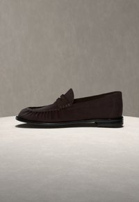 Mocassin en cuir marron foncé avec une bride penny classique, un petit talon et des coutures de style mocassin sur une surface claire.