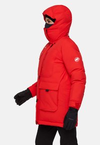 Parka isolato rossa con cappuccio, multiple tasche frontali e una zip nera. Guanti neri completano l'outfit per il calore.