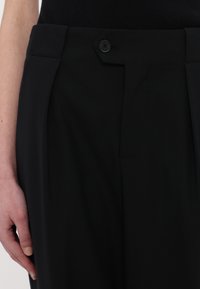 Musta räätälinhousut, joissa on sileä pinta, yksinkertainen napitus ja etupleatit rakenteellista muotoilua varten.