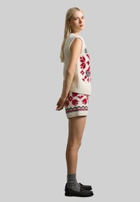 Häkelter ärmelloser Pullover und Shorts-Set in Creme, mit roten Blumenmustern und schwarzen Akzenten. Kombiniert mit grauen Socken und schwarzen Schuhen.