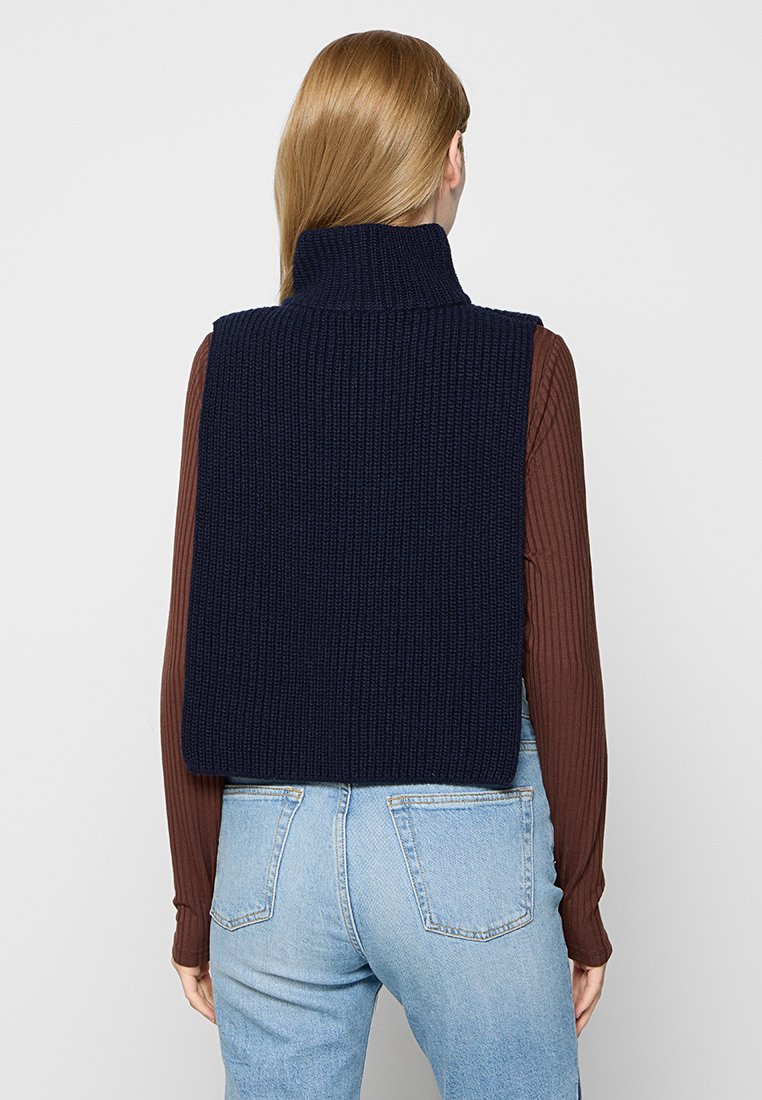 Gilet en tricot côtelé marine avec col haut, porté sur une chemise à manches longues marron, associé à un jean bleu clair. Coutures visibles sur le jean.
