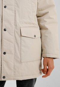 Chaqueta aislante beige con textura suave, gran bolsillo frontal con cierre de presión y detalles de herrajes plateados.