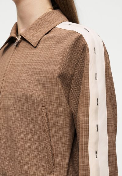 Κοντινό ενός καφέ καρώ σακακιού με φερμουάρ, γιακά, πλαϊνή τσέπη και άσπρη ρίγα στο μανίκι με μαύρα λογότυπα.